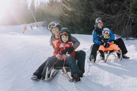 Mutter und Kind sowie Vater und Kind sitzend auf je einer Rodel