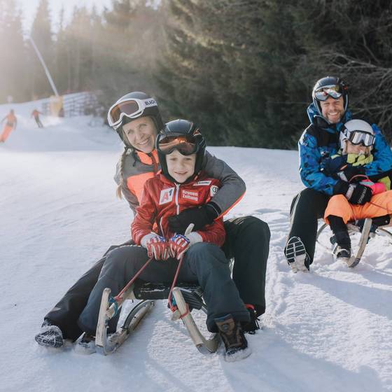 Mutter und Kind sowie Vater und Kind sitzend auf je einer Rodel